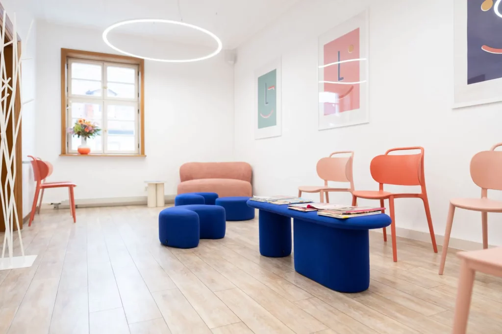 Wartezimmer mit Bildern an der Wand, rosane Stühle, rosanes Sofa, einem blauen Tisch und blauen Hockern